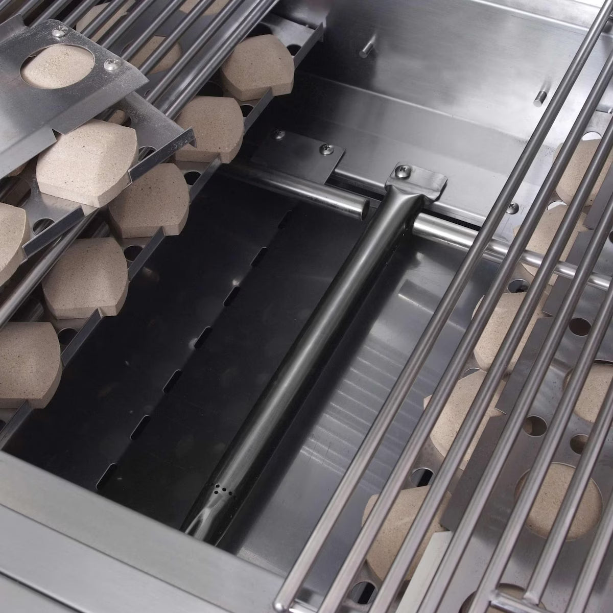 Close-up inside a Renaissance Cooking Systems grill firebox showing a tubular stainless steel burner running along the floor, flanked by ceramic briquettes in their trays beneath the cooking grates