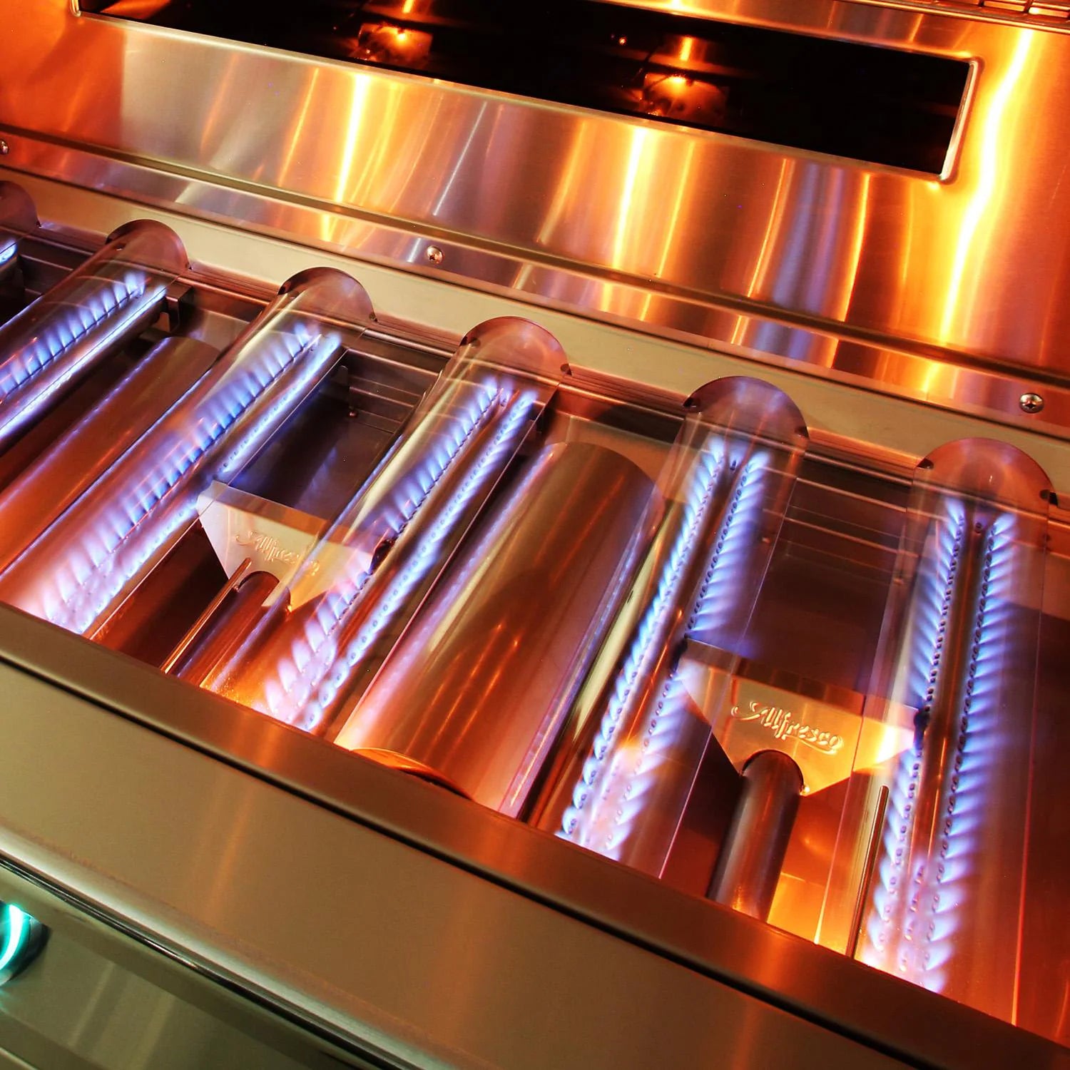Close-up of a lit Alfresco grill firebox interior showing rows of stainless steel tube burners with blue gas flames, the Alfresco logo visible on the burner shields
