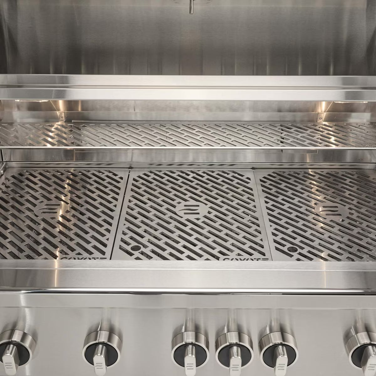 Front interior view of an open Coyote stainless steel grill showing the cooking grates, the warming rack above and a row of control knobs along the bottom