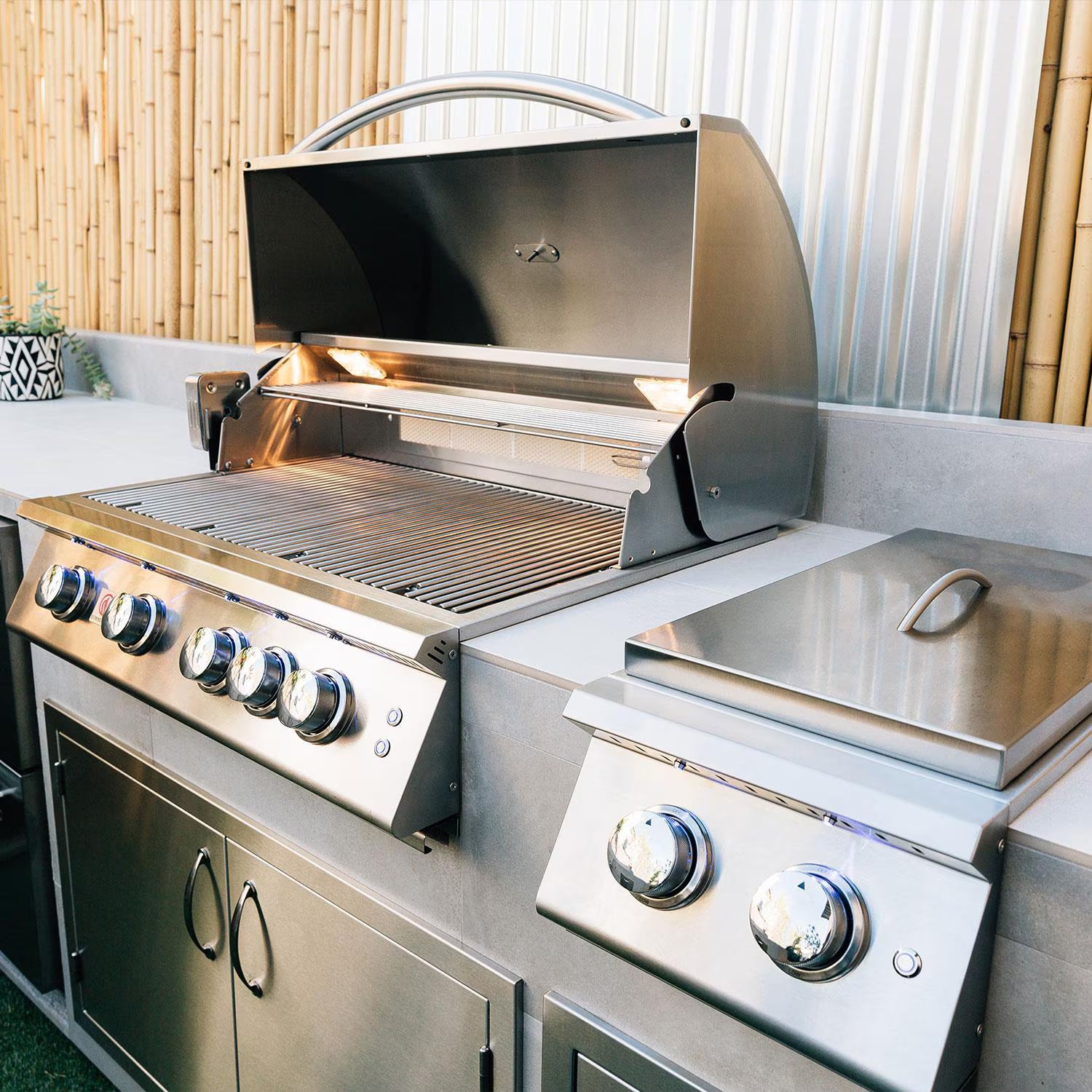 Summerset built-in gas grill with hood open beside a side burner in a stainless steel outdoor kitchen island