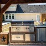 A lifestyle thumbnail of an outdoor kitchen island with a built-in stainless steel gas grill in a residential backyard, with a house in the background