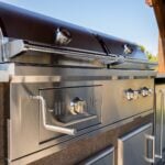 A close-up detail thumbnail of a stainless steel built-in gas grill showing the upper barrel hood and control area at an angle