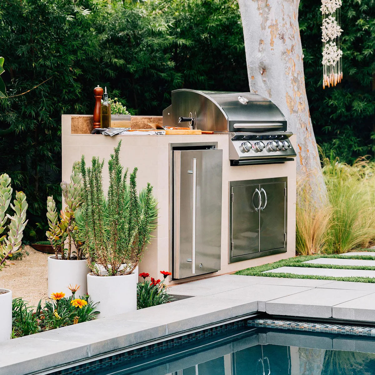 Summerset built-in gas grill and refrigerator in a stucco outdoor kitchen island beside a pool with landscaped plants