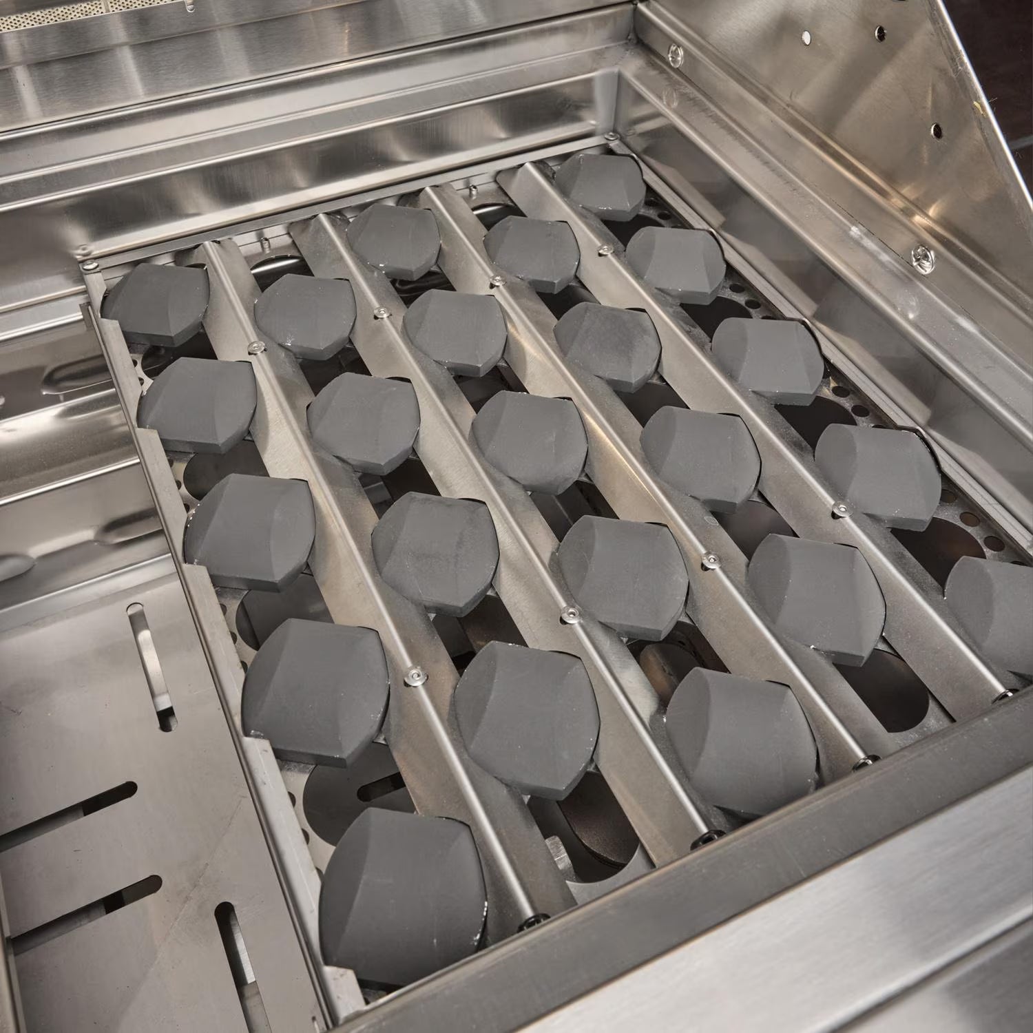 Angled interior view of a grill firebox showing rows of dark ceramic radiant briquettes seated in metal channels above the burners