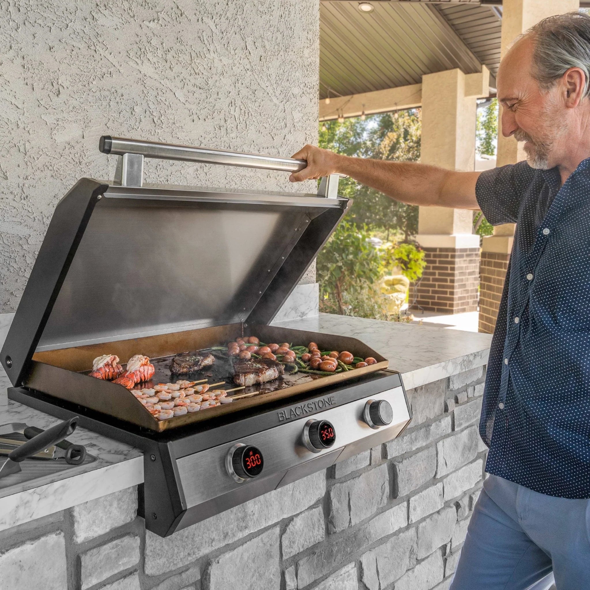 Man opening the lid of a Blackstone 30-inch electric drop-in griddle built into a stone outdoor kitchen island while food cooks