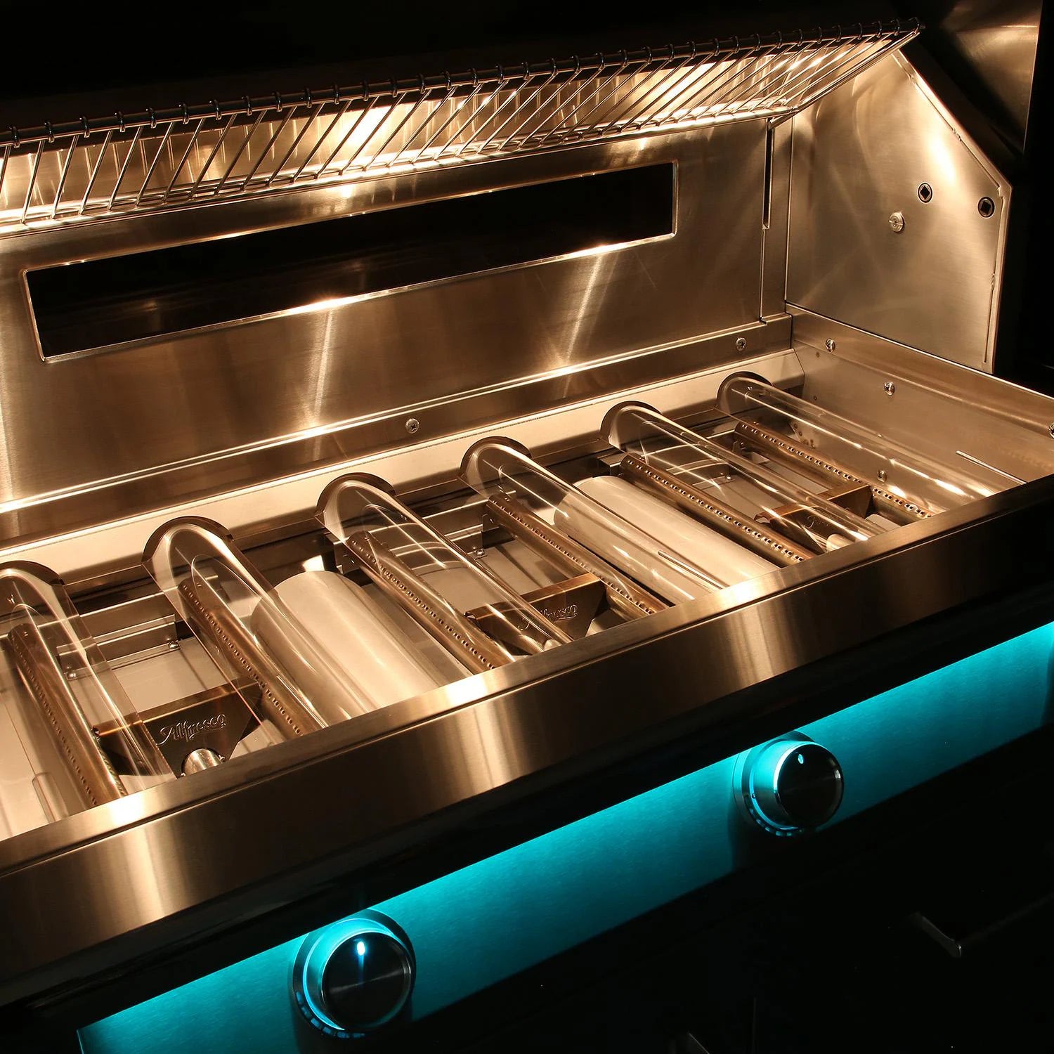 Angled interior view of an open Alfresco stainless steel grill lit by its interior lights, showing the tube burners and warming rack, with control knobs glowing teal-blue on the front panel