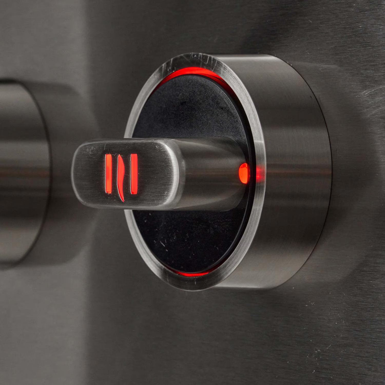 Close-up of a stainless steel grill control knob with red illuminated backlit position markings and a glowing red ring around its base, on a brushed metal panel