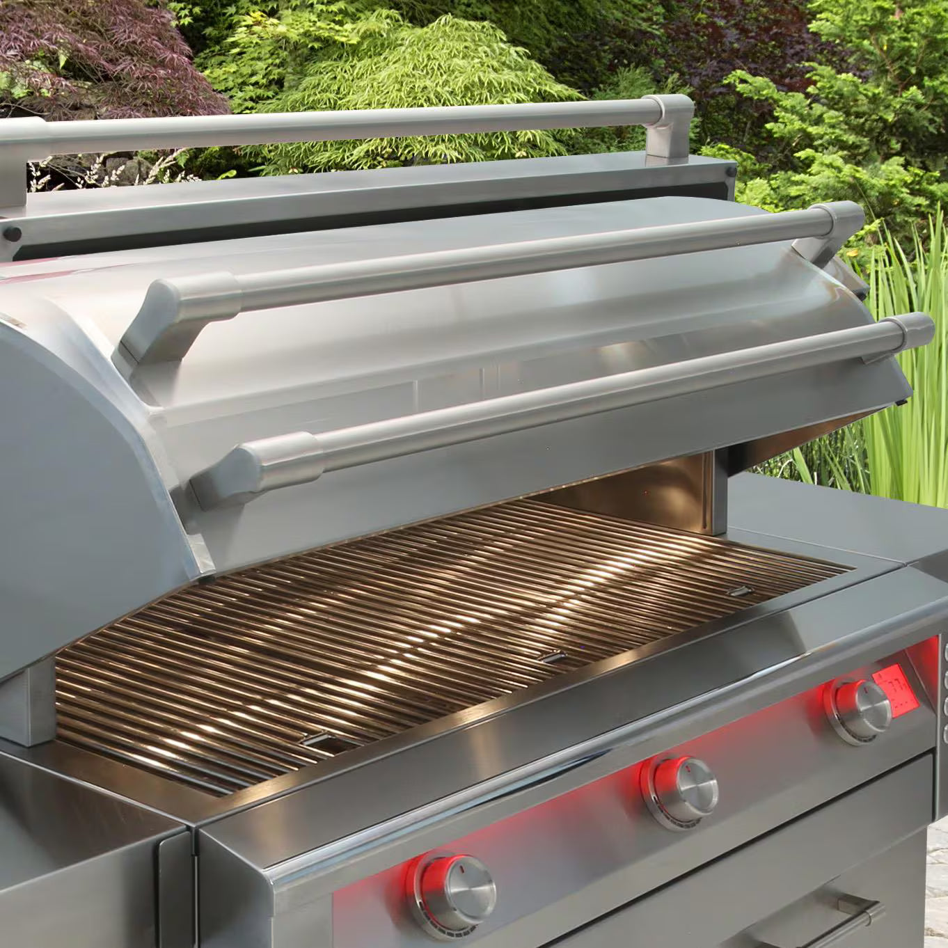 Built-in stainless steel grill with the lid open, showing the cooking grates and a row of red-backlit control knobs, set in a lush green outdoor garden