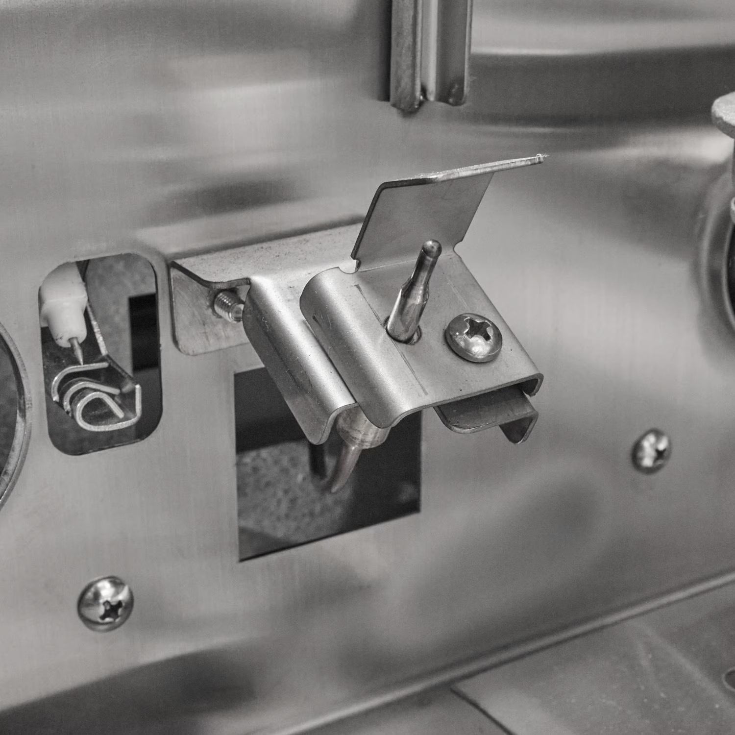 Close-up inside a grill firebox showing the ceramic flame-thrower ignition electrode and a stainless steel burner mounting bracket near a burner port