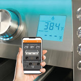 A hand holding a smartphone running a grill-monitoring app showing grill zone temperatures and a wireless food probe reading, in front of a stainless steel grill control panel with a blue LCD temperature display and backlit knobs