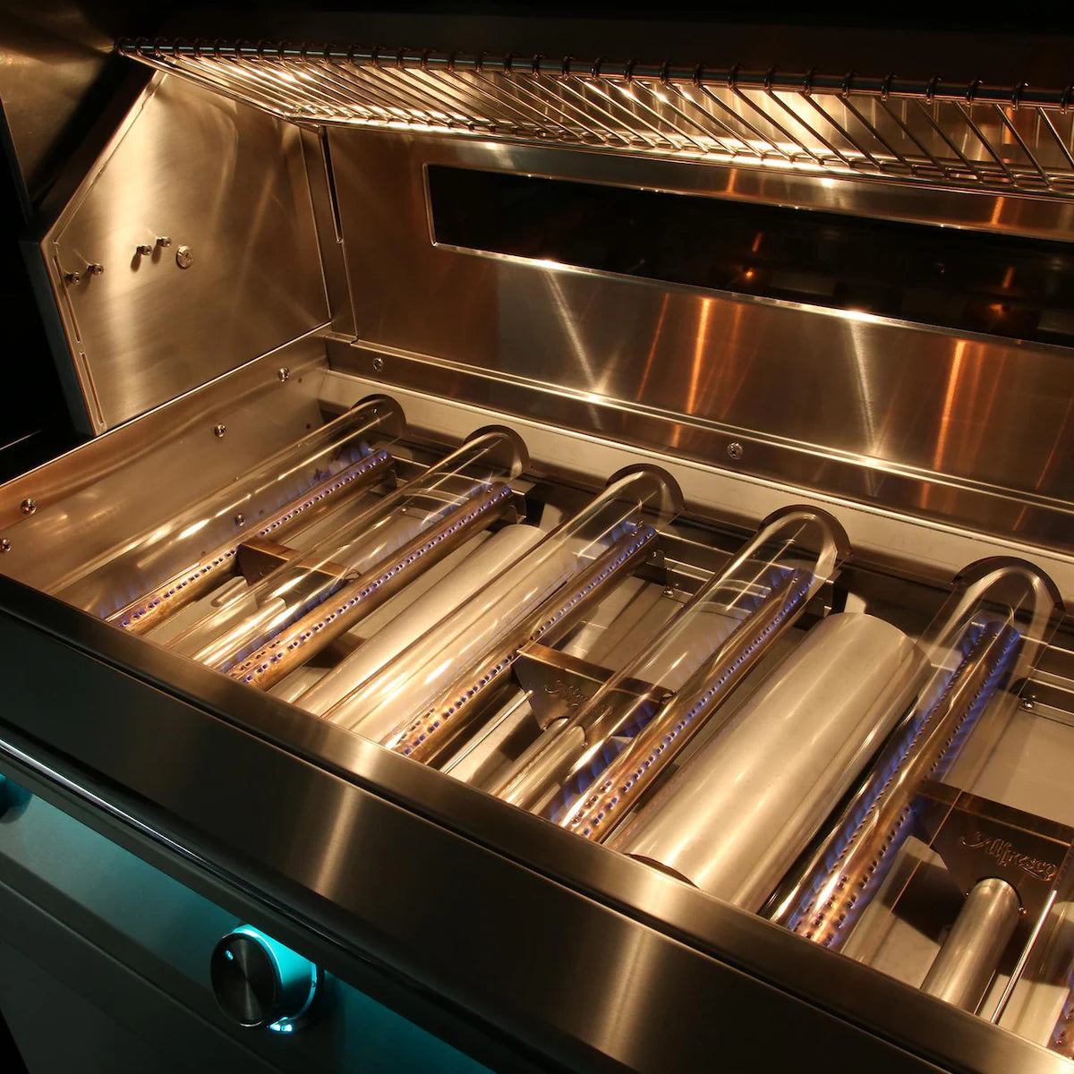 Interior of an open Alfresco built-in grill with the lid raised, showing the rows of stainless steel tube burners and grates removed, the warming rack above, and a teal backlit control knob