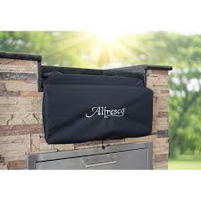 Black Alfresco vinyl cover with the white Alfresco script fitted over a burner set into an outdoor kitchen counter, photographed outdoors with sunlight behind