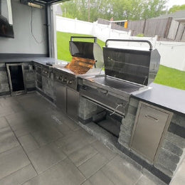 Large stainless steel built-in outdoor kitchen with a gas grill and stone-veneer cabinets on a tiled patio in a fenced backyard
