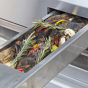 Stainless steel smoker tray filled with wood chips and herbs inside an Alfresco built-in gas grill