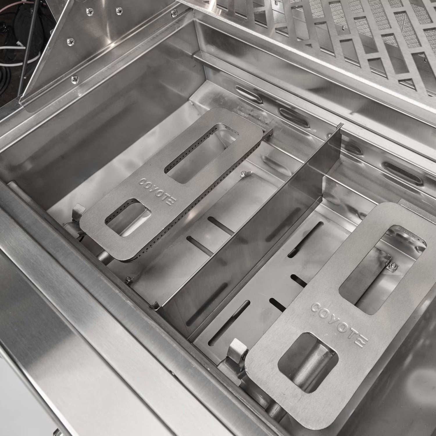 Interior of an open Coyote grill firebox showing the Coyote-branded stainless steel heat diffuser plates seated over the burner tray, with the warming rack above
