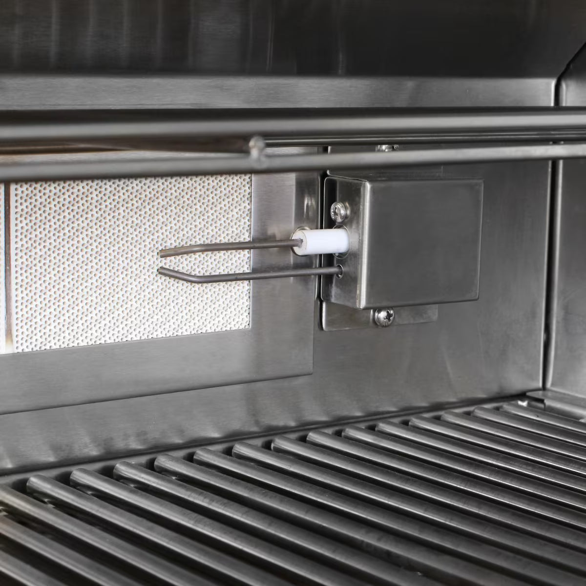Close-up inside a grill firebox showing the rear infrared burner with its mesh screen, the ceramic ignition electrode and a metal junction box mounted at the back, above the cooking grates