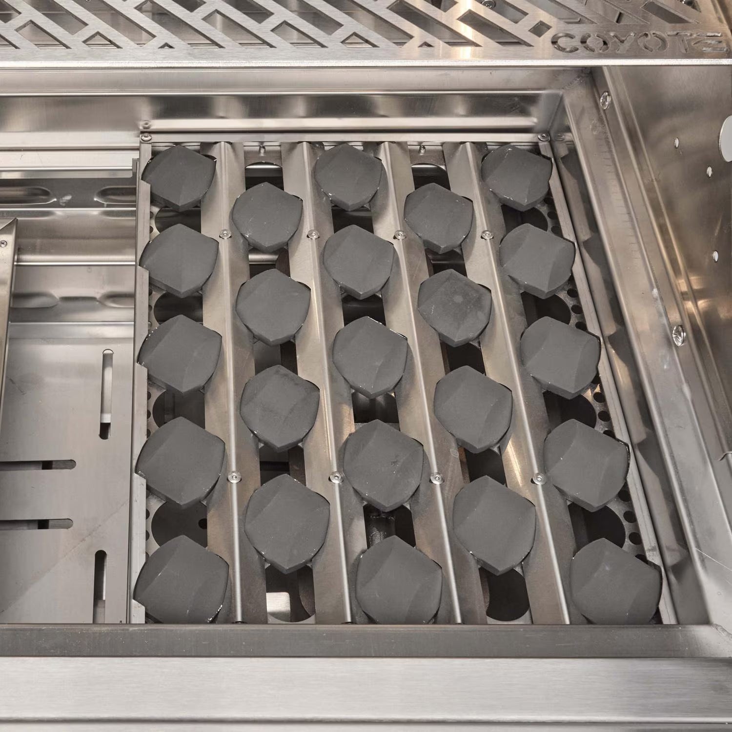 Interior of a Coyote grill firebox showing rows of dark ceramic radiant briquettes seated in metal channels above the burners, with the Coyote-branded warming rack at the top