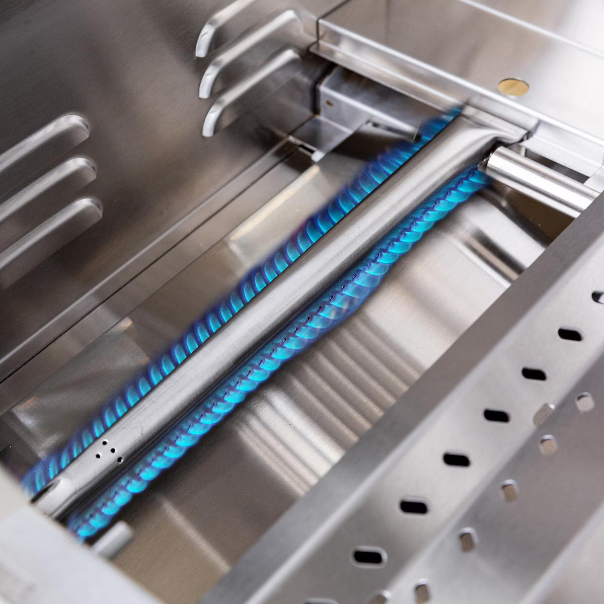 Close-up of a Summerset Sizzler grill interior showing a lit stainless steel tube burner with blue flames and the warming rack above