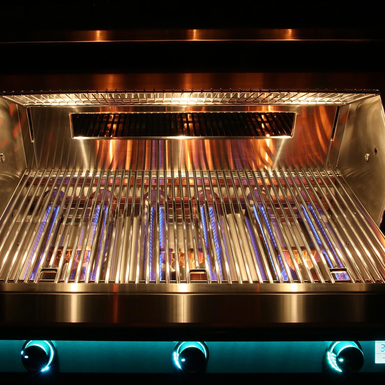 Interior of a lit built-in stainless steel grill with the lid open, showing blue gas flames beneath the cooking grates, a warming rack above, and three teal-blue backlit control knobs along the bottom