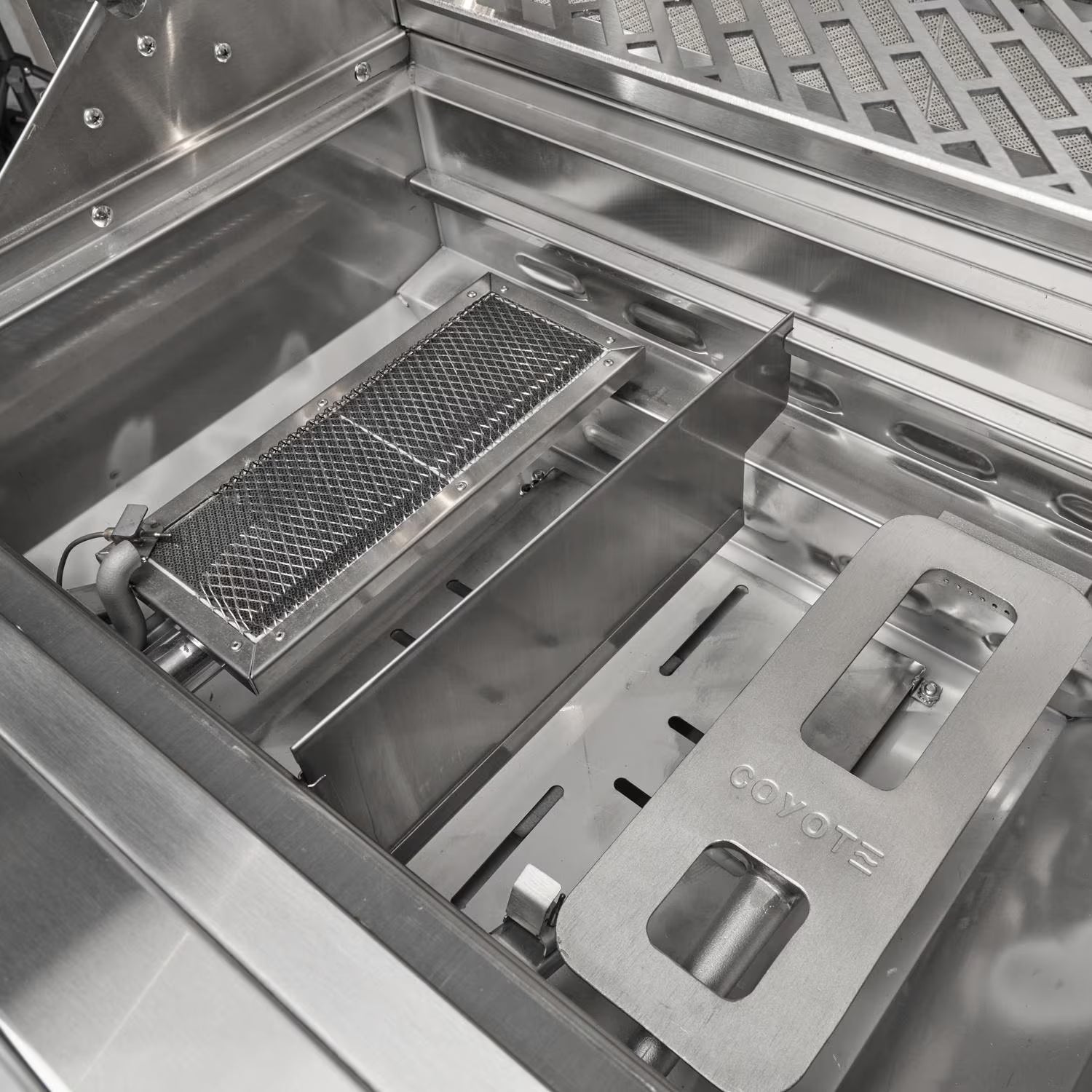 Interior of an open Coyote grill firebox showing the infrared sear burner with its mesh screen beside a Coyote-branded heat diffuser plate, over the burner tray