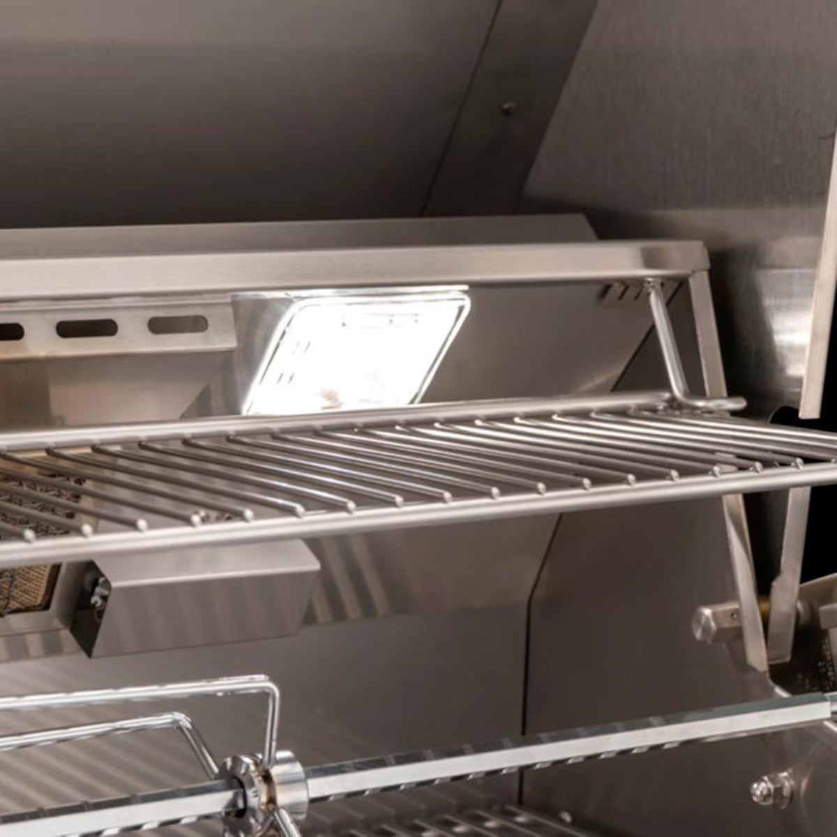 Interior of an open American Outdoor Grill showing stainless steel cooking grates, warming rack, halogen interior light, and rotisserie rod