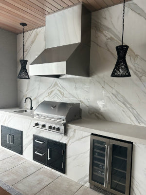 Luxury outdoor kitchen with marble backsplash, stainless steel range hood, built-in gas grill, black cabinetry, and glass-door beverage fridge