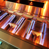 Close-up of a lit Alfresco grill firebox interior showing rows of stainless steel tube burners with blue gas flames, the Alfresco logo visible on the burner shields