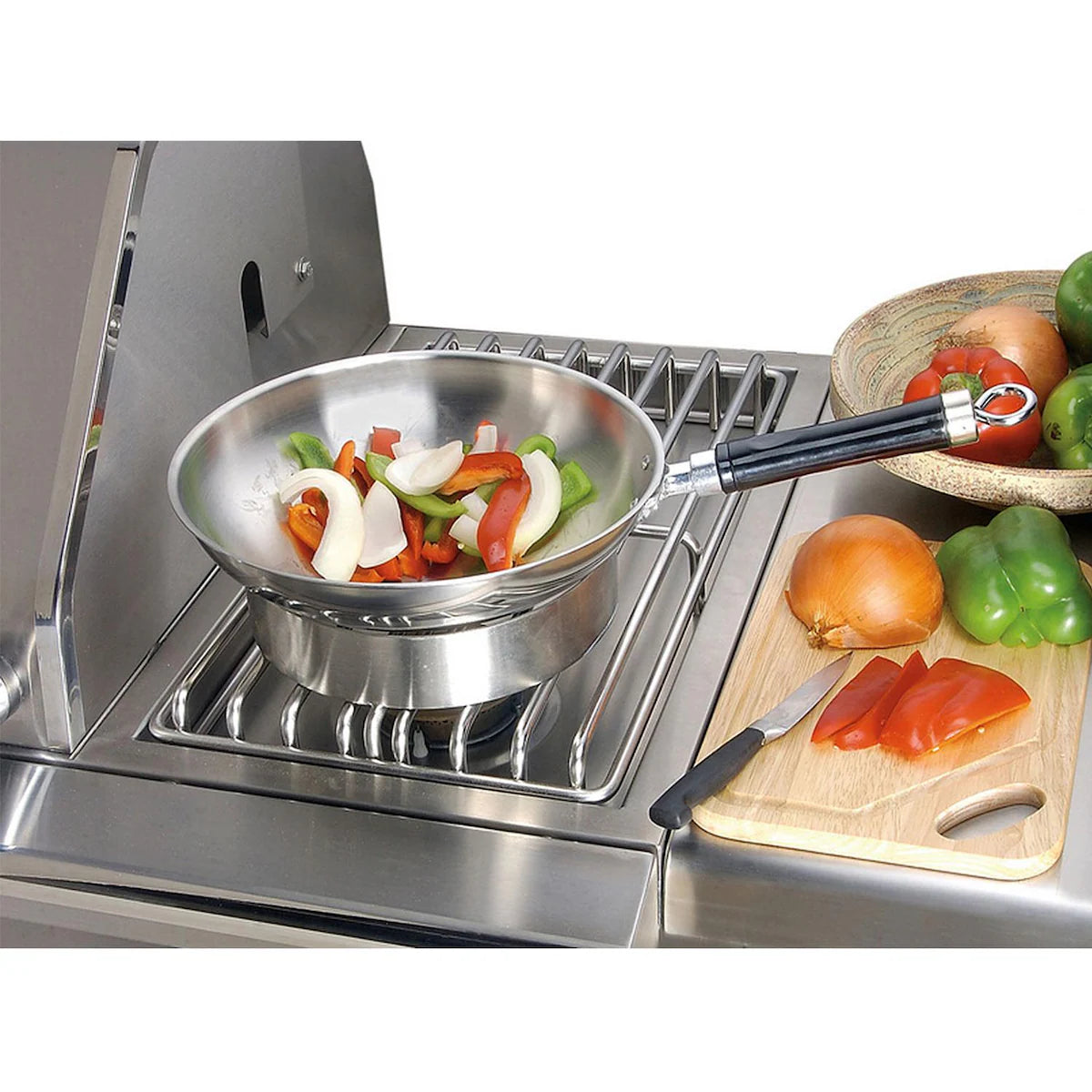 Alfresco dual side burner set flush into a stainless steel surface beside a grill, with a saute pan of sliced peppers and onions cooking on the burner and a wooden cutting board of vegetables alongside