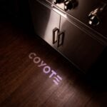 A dimly lit feature thumbnail showing the Coyote logo projected in light onto a wooden floor beneath a grill, demonstrating the illuminated control knob throw-light feature