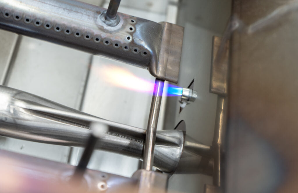 Close-up of AOG's Flame Thrower burner igniting on the grill's gas manifold, a product feature shot