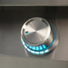 Close-up of a brushed stainless steel grill control knob with a teal-blue backlit LED ring and illuminated indicator markings, on a stainless steel panel