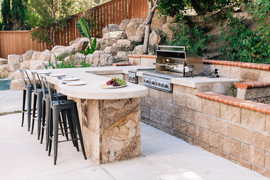 Summerset built-in gas grill in a stone outdoor kitchen island with a curved bar counter and stools in a landscaped backyard